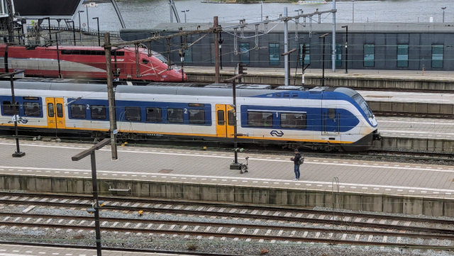 A railway station with several platforms and two trains. On an otherwise empty platform, there is a semi folded Brompton bicycle and a man taking picture of it.