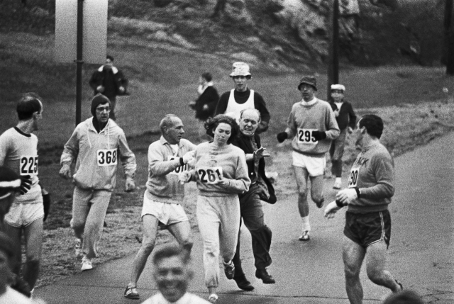 Katherine Switzer running along a road in a grey tracksuit. She is a white woman with dark hair. There are several other men running alongside her, also in running gear. They are protecting her from a man in a blazer who is attempting to rip off the number pinned to her back.