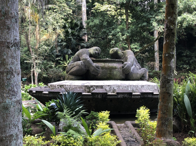 Foto de una escultura sobre una fuente, en medio de un parque repleto de vegetación. Son dos personas abrazando una gran cazuela, talladas en piedra. Tienen manos gruesas y poderosas.