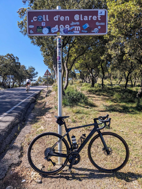 A black carbon road bike leaning against a road sign that reads: Coll d'en Claret. 498m (of elevation). The sign is full of various stickers so it's almost hard to read. It's missing a Conversations sticker though... 