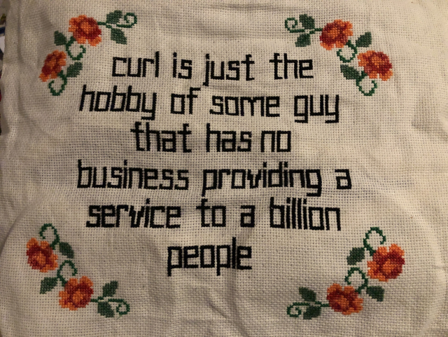 Pillow with white Aida-cloth. Cross-stitched roses at the corners, text: “curl is just the hobby of some guy that has no business providing a service to a billion people”
