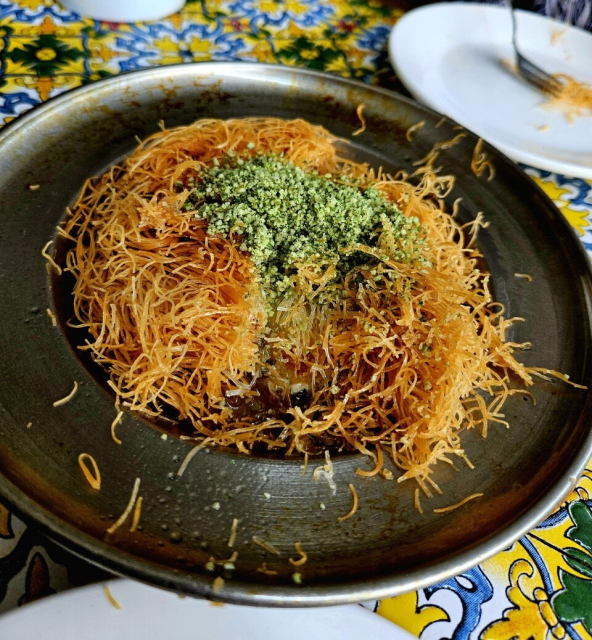 A copper plate of golden kunafa with a generous mound of crushed pistachios on top. It's already by cut open to reveal the centre. The crispy flour noodles absolutely dominate the dish though you can still see some of the melted cheese in the middle.