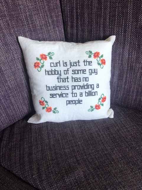 Same pillow as above (red roses bordering “curl is just the hobby of some guy that has no business providing a service to a billion people”), but now properly stuffed. Resting on a lilac couch 