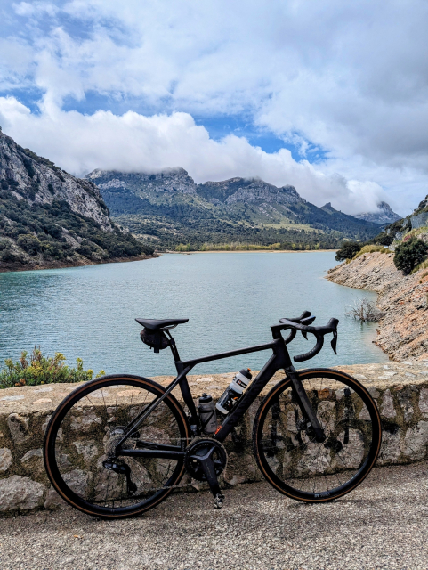 A black carbon road bike leaning against a low stone wall in front of lake with mountains in the background.
