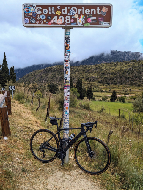 A black carbon road bike leaning against a road sign that reads "Coll d'Orient". A grassy green landscape behind. So mountains in the background are obscured by low hanging clouds. 
