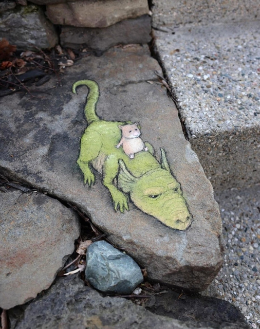 Streetart. A dragon with a hamster have been painted with chalk on a triangular gray stone slab. The small beige hamster is sitting on the back of the reclining dragon with its eyes closed and chilling.
Title: "Angharad is a fierce and solitary hunter, but sometimes her back gets itchy."