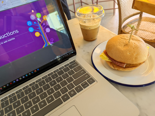 Looking over a marble cafe table, my laptop open with a purple slide and AccessU logo of a stylized tree visible, an iced coffee drink and breakfast sandwich (egg, cheese, prosciutto, GF roll) sitting alongside.