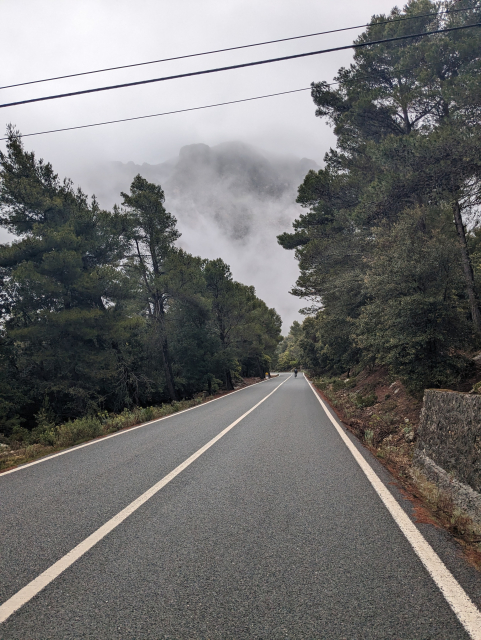An lonely road going up a mountain. In a distance another cyclist going up the road. The mountain in the background is barely visible through the clouds.