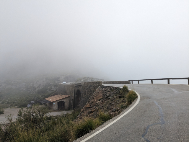 The famous 270° corner of the road down to Sa Colabra. The road passes underneath itself. The background is entirely covered by low hanging clouds. 