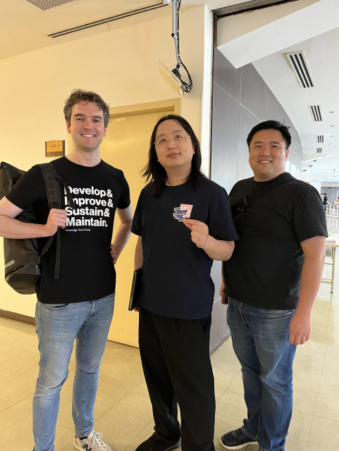 Paul Sharratt (wearing a Develop& Improve& Sustain& Maintain t-shirt), Audrey Tang (holding a "move deliberately and maintain things sticker) and Powen Shiah in a hallway at g0v summit