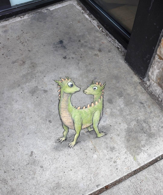 Streetart. In front of a store, two green dragons have been painted with chalk on a rectangular sidewalk slab. The two appear to be Siamese twins, as they only have one body but two heads. The two look at each other. Title: "Herbert and Helen wrestle with the awkward realization that neither one of them is the better half."