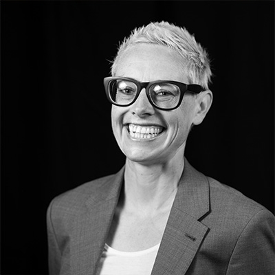 Black and white photo of Anna Zivarts. Anna has short, light colored hair and black rimmed glasses. She is smiling and wearing a grey blazer and white shirt.