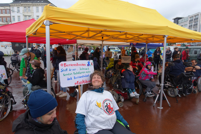 Demo mit vielen Menschen im Rollstuhl - aber nicht nur. Es ist nass, viele Menschen stehen unter Pavillonzelte. ich sitze auf meinem Rollstuhl mit T-Shirt mit einem roten und schwarzen rollstuhlfahrenden Menschen - im Still wie im Antifa Logo. und ich trage ein Plakat "#BarriereBrechen Stell dir vor .... Jederzeit spontan Bahnfahren möglich dank Barrierefreiheit #FightAbleism"