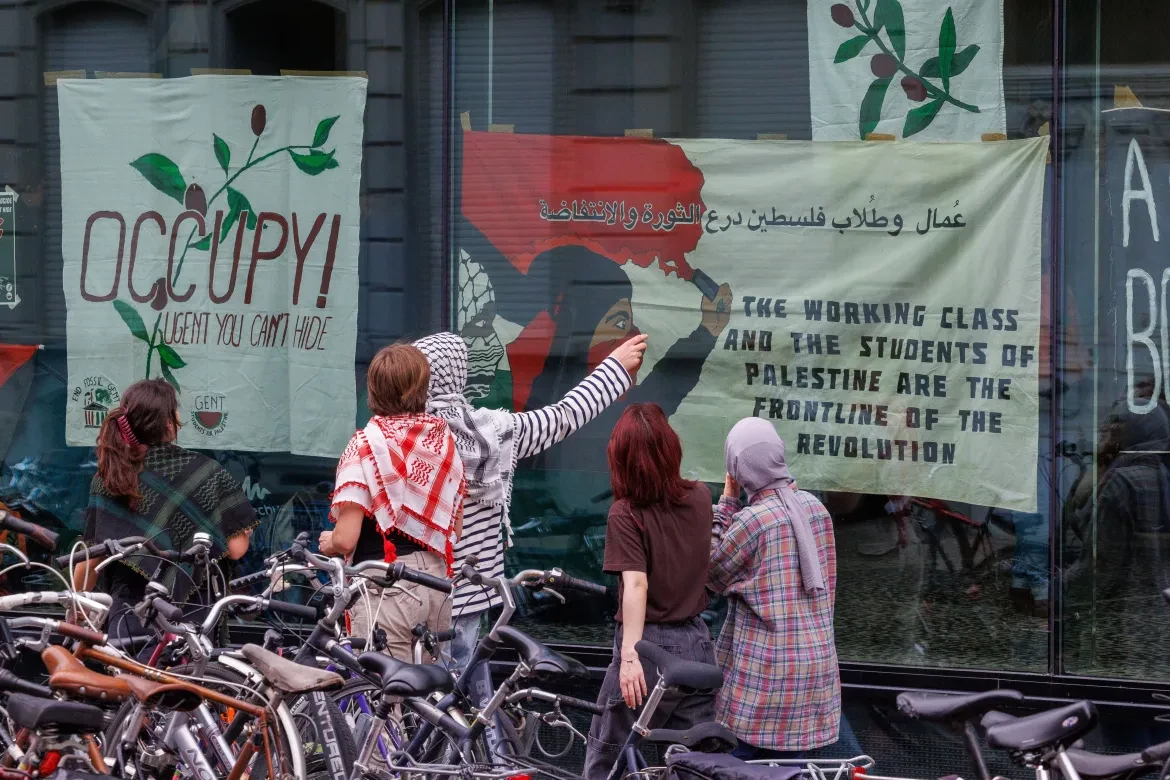 Fotos van de ramen van het Universiteitsgebouw. Er hangen banners aan: eentje met 'Occupy, UGent, you can't hide', de andere met een Moslima die een rookbom vasthoudt en "The working class and the students of palestine are the frontline of the revolution"

Enkele studenten staan buiten en kijken naar de banners.