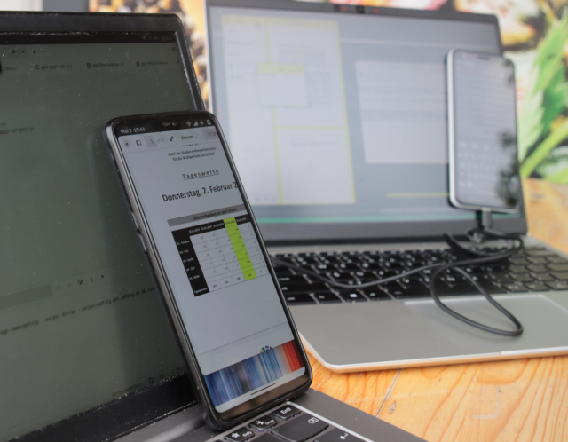 Two notebooks running GNOME – Builder on Debian on the left, Papers on postmarketOS on the right – with each having a postmarketOS running phone (OnePlus 6 and Google Pixel 3a) leaning against the notebooks' displays.