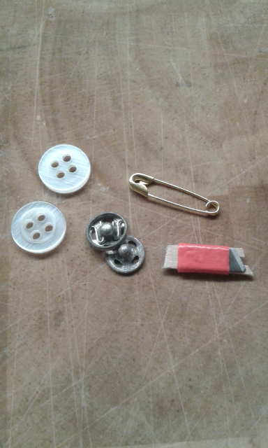 Two tiny shirt buttons, a press-stud, a safety pin and a cutter made from a snap off craft knife blade taped to a scrap of card with a notch cut to expose the middle of the blade. 