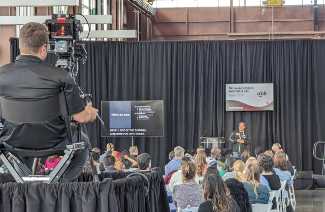 Man speaking on stage. There is a monitor behind him and next to him. People sitting in the audience. A camera man is facing the stage in the foreground.