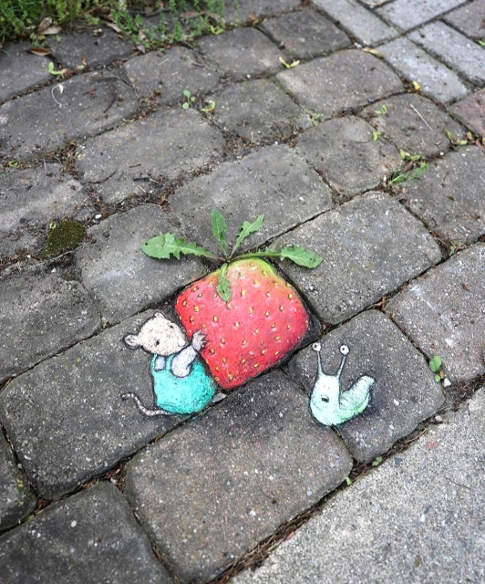 Streetart. A mouse with a strawberry and a snail was drawn with chalk on a gray sidewalk made of cobblestones. A little mouse (called Fiona) in a turquoise dress is holding a large square strawberry, which is being admired by a green snail next to it. She is standing in front of the strawberry with her mouth open. The strawberry was painted on a square paving stone, green weeds in the crack serve as strawberry stalks. Title: "Fiona goes to great lengths to keep her heirloom strawberries away from the slugs."
