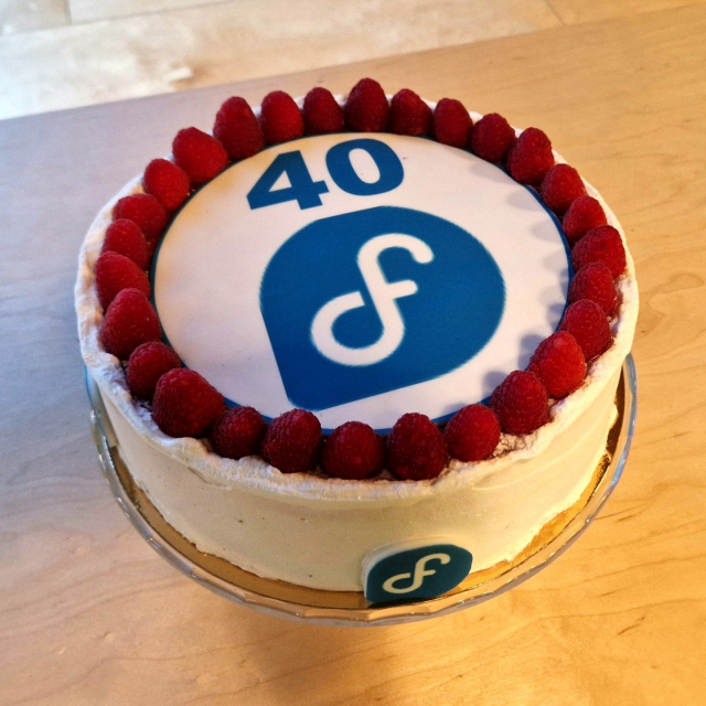 A white cake that has the Fedora Linux logo and the number 40 on the top. There is a ring of raspberries.