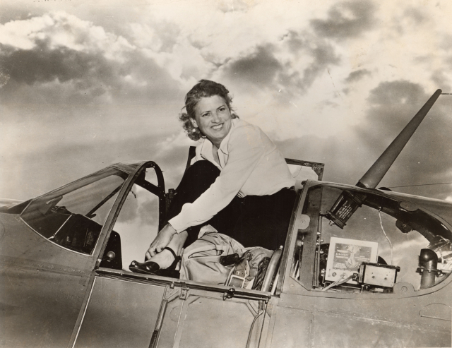 A posed photo of Jackie Cochran sitting on the edge of her plane. She's a white woman with blonde hair and is rather implausibly wearing high heels. Breaking the sound barrier as a pilot? In these shoes? I don't think so.