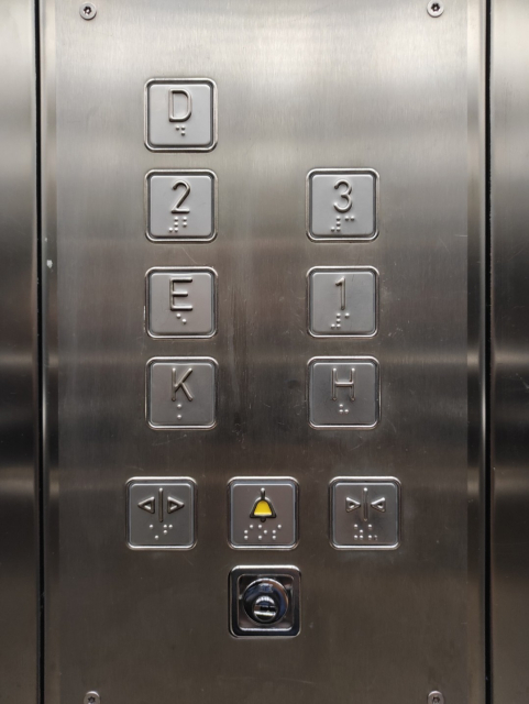 10 Tasten und ein Schließzylinder eines Lifts aus Edelstahl. Die Tasten sind mit fühlbarer Profilschrift und mit Braille beschriftet. Auf den Tasten steht in zwei Spalten „D“, „2“, „E“, „K“, „3“, „1“, „H“ und in einer Zeile darunter steht „auf“, „sos“ und „zu“. Am oberen und unteren Rand des Fotos befinden sich Schrauben mit Sicherheits-Torx Antrieb in der Edelstahlvertäfelung.