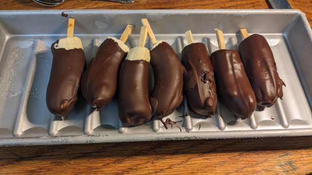 A row of banana halves with chop sticks in them dipped in chocolate sit on a hotdog bun pan.