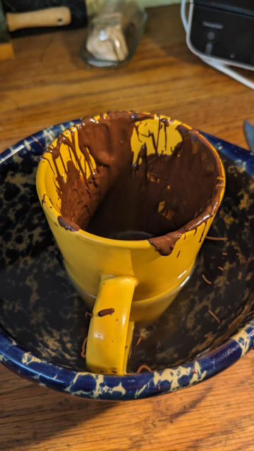 A tall yellow cup with a mess of melted chocolate inside it sits in a speckled blue Bennington potters dough mixing bowl in hot water.