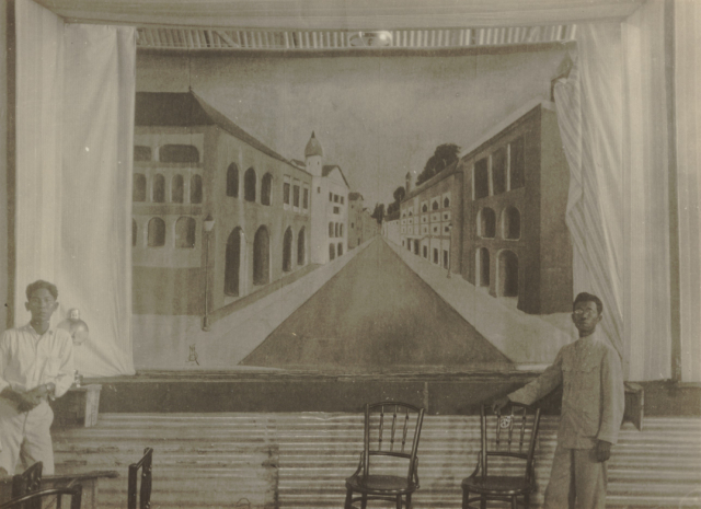 black and white photo of two Indonesian men standing in front of a painted mural of an urban street scene. there are curtains on either side and chairs scattered around in the foreground.