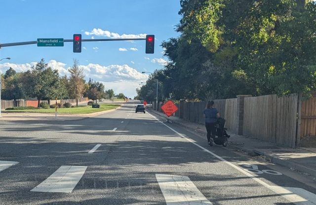 A photo of a road with the bike path blocked off by a "left lane closed ahead" street sign. The picture is taken from a bit of a distance, with a car visible driving further away from the viewer, and a person with a baby carriage crossing the street.