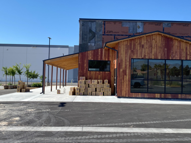 A building under construction with wooden and metal exterior. Stacks of Playdate cartons are visible outside near the entrance. The surrounding area is paved with a newly constructed sidewalk and some young trees in planters. The Playdate boxes are hastily strewn about on the sidewalk.