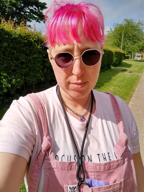 Alexis, a woman with light skin tone and bright pink hair shaved at the sides and tied back, a fringe on her face. She is wearing pink rimmed glasses which have tinted in the sun, a pink t-shirt and pink cord dungarees. She is outside on a sunny day.