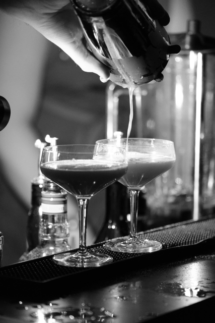Schwazweiß-Foto von zwei Cocktailgläsern an einer Bar die gerade aus einem Shaker befüllt werden.