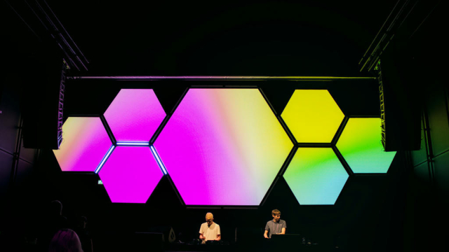 Bühne mit zwei DJs auf. Im Hintergrund sind mehrere Hexagone mit bunten Farben zu sehen.
