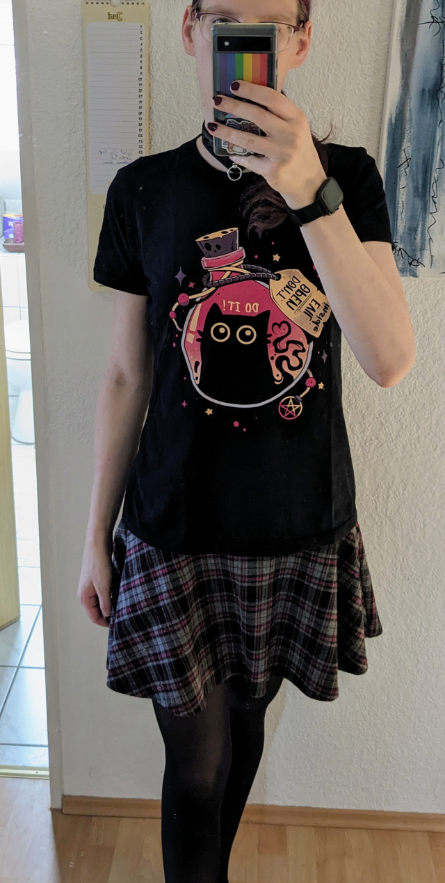 Foto von mir, in Strumpfhosen, kariertem Rock, schwarzem T-Shirt mit nerdigem Motiv, Halsband, und dem Gesicht as usual verdeckt von meinem durchaus bunten Smartphone 