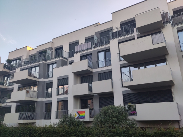 A modern multi-story residential building with a progress pride flag hanging on one of the balconies.