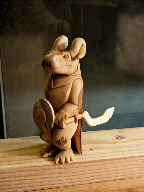 Woodcarved kobold wearing a hooded cape, left hand on his scimitar-like pommel, wielding a shield by his right hand. Its horns are curved, a bit like ram horns do.