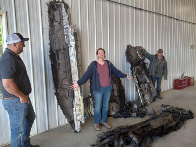 Me standing amidst a collection of partly burned metal and carbon fibre space junk leaning on a white metal wall.  I'm looking at the camera with my arms outstretched and with a yelly-smiley face.  The piece of junk next to me is much, much taller than I am.  There are two farmers in the background who are the ones who found pieces of junk on their land.