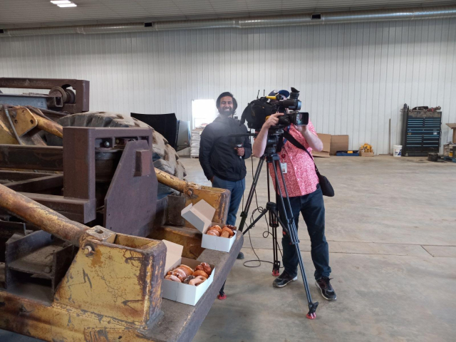 2 boxes of donuts on the back of a very heavy duty truck thing, inside a big open shop.  A reporter is smiling at my camera, while a reporter next to him sets up a TV camera.