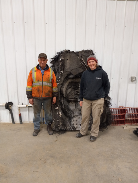 Me wearing a black hoodie and grey pants, standing next to a farmer wearing an orange reflective jacket and jeans.  We're both looking at the camera and smiling.  Behind both of us is an enormous piece of space junk - it's 6 feet tall and 4 feet wide, with frayed, burned up edges.  It's leaning on a white metal wall.