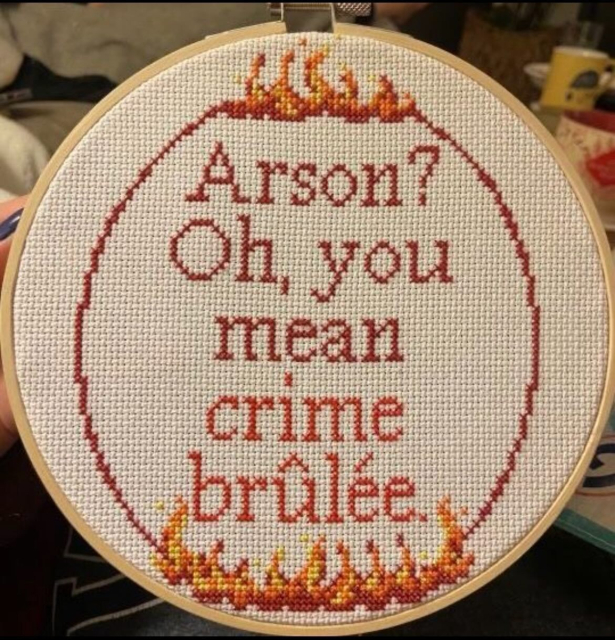 A round cross-stitch embroidery in a wooden hoop with the text 'Arson? Oh, you mean crime brûlée.' The text is stitched in red, and the top and bottom edges of the hoop are decorated with small stitched flames in red, orange, and yellow.