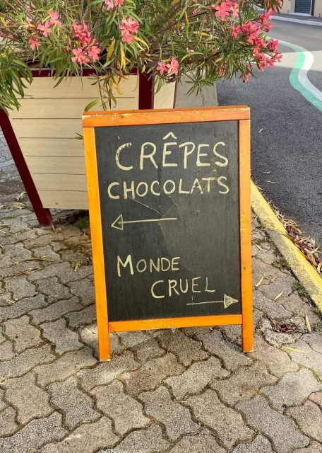 Small sandwich-type sign on a French street. Arrow pointing to the left says "crêpes chocolats".  Arrow pointing right reads "Monde cruel" (cruel world) 