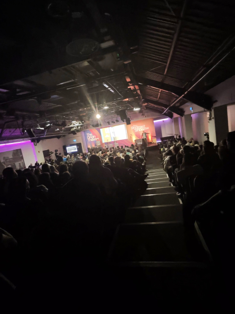 The image shows a large, dimly lit auditorium filled with people seated and facing a stage. The stage is brightly lit with spotlights, and there are large screens or banners on the stage with the text "The Origin Theatre" and "The Podcast Show 2023." The audience appears to be attentively watching a presentation or panel discussion. The ceiling has an industrial look with exposed beams and lighting fixtures. There is a central aisle leading down to the stage, and some people in the audience are holding up their phones, possibly taking pictures or recording the event.