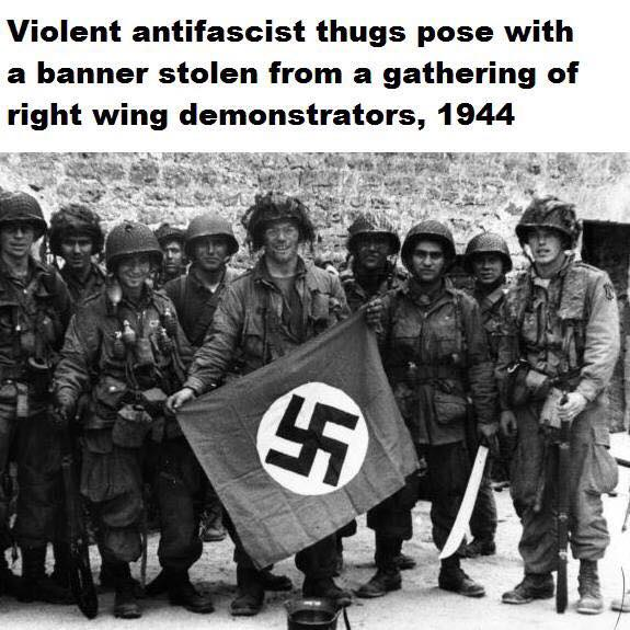 B&W photo of US Airborne soldiers in WW2 posing with a captured Nazi flag. Caption reads "Violent antifascist thugs pose with a banner stolen from a gathering of right wing demonstrators, 1944"