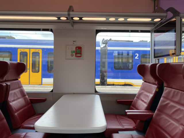 Interior of a train. A bay of four with red leather seats and a big table. The wall between windows is in the middle of the bay but not really so that the table is off centre.