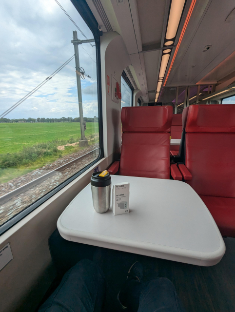 A view from a train seat. There are two red seats in front, full window view on the left and some ceiling LED lights.
On the table there's a Thermo mug from IKEA as well as a paper box of 1x SJÖSS 2x USB-C PD (45W) charger.