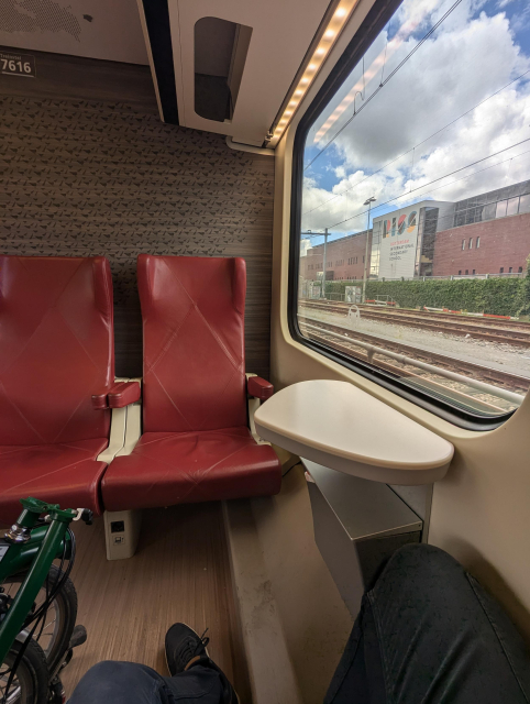 A view from a train seat. There are red seats across and a window on the right. The floor goes up underneath the right seat. A part of green folded bicycle is visible on the left.