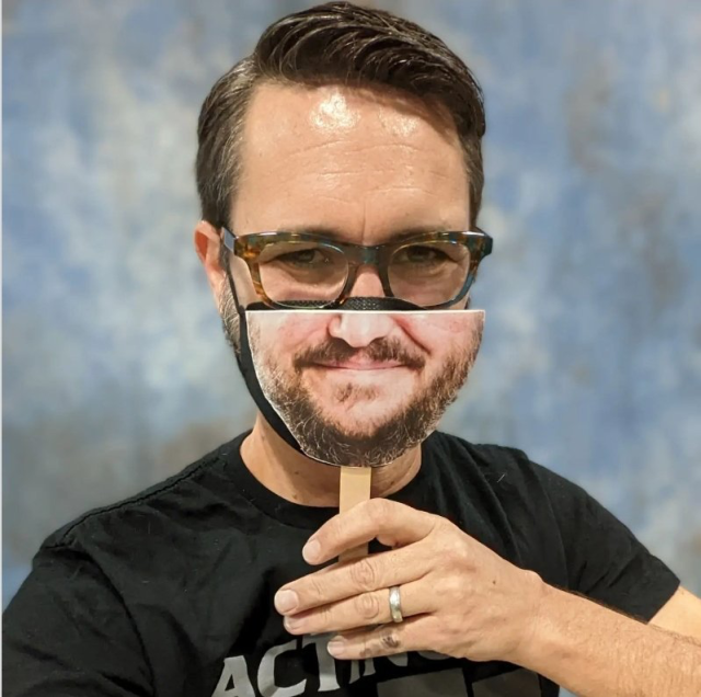 Wil Wheaton is wearing horn-rimmed glasses, a black respirator and is holding up a cardboard cutout of the lower half of his face on a tongue depressor up over his respirator.