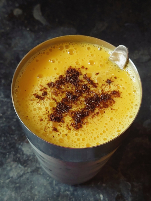 On a kitchen counter of black stone, a tall steel tumbler with a steel spoon peeking out, 98% full of fresh mango lassi made from homemade curd and Himsagôr mangoes. It's brilliant golden yellow in colour, a bit foamy on top from stirring some water in (for the correct smooth but not cloyingly heavy consistency), and visibly intense in texture. A judicious amount of homemade bhaja môshla (traditional Bengali spice blend) has been streaked in on the top, that has moistened and turned an inviting almost-coffee brown but with a distinguishing red undertone.