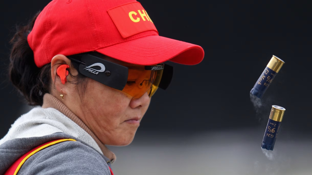 An older Zhang Shan competing in a red China baseball hat, with 2 ejected shotgun shells, smoke curling from their ends in 2012
