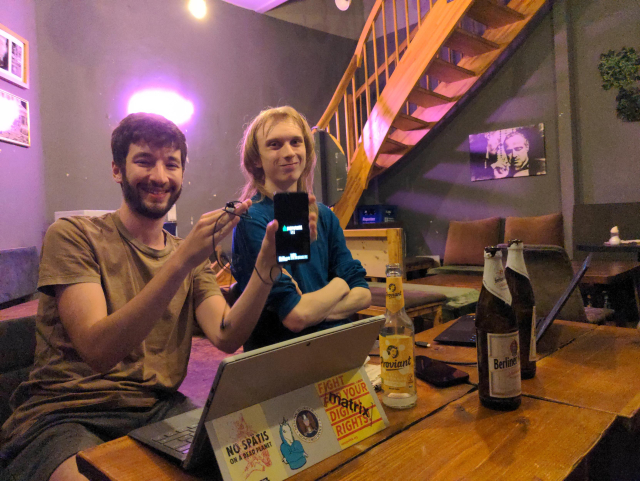 Jonas and Caleb sitting at a table in a cafe at 1AM with their laptops and some drinks. Jonas is holding a phone with the postmarket bootup screen and some USB headphones.